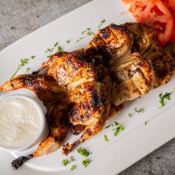 Grilled whole chicken with charred skin, served with a white sauce and sliced tomatoes on a white plate.