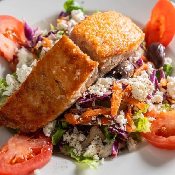 Golden-brown salmon fillets on a fresh salad with crumbled feta, tomatoes, olives, and shredded carrots.